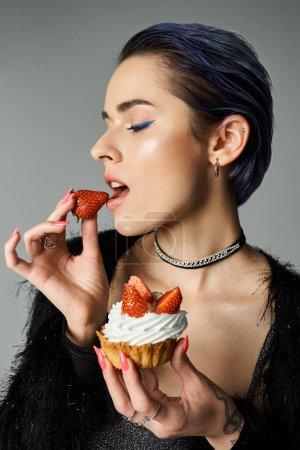 A young woman with vibrant blue hair enjoys a cupcake in a stylish studio setting.