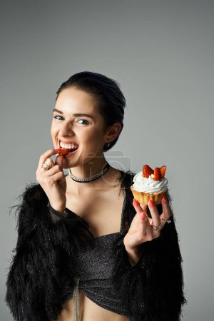 Stylish woman with short dyed hair enjoys a cupcake in a black attire, celebrating a special occasion.