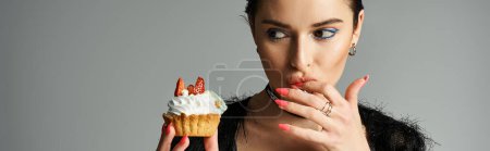 Téléchargez les photos : A young woman with dyed hair holding a cupcake in her right hand, exuding joy and celebration in stylish attire. - en image libre de droit