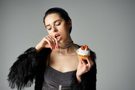 A stylish woman with short dyed hair poses while delicately holding a cupcake in a studio setting.