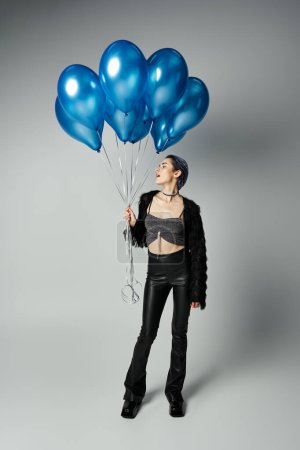 A young woman with short dyed hair and stylish attire joyfully holds a bunch of blue balloons in a studio setting.