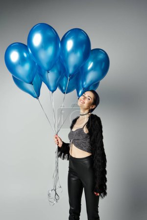 A stylish young woman with short dyed hair holding a bouquet of blue balloons in a studio setting.