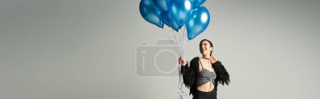 A woman with short dyed hair looks festive while holding a bunch of blue balloons in a studio setting.
