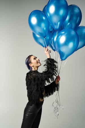 A young woman with short dyed hair poses in stylish attire, holding a bunch of blue balloons.