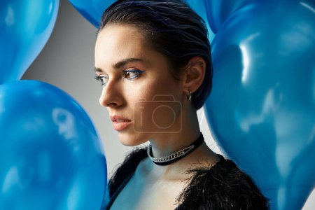 A stylish young woman in a black dress gracefully holds a cluster of blue balloons, exuding elegance and joy.