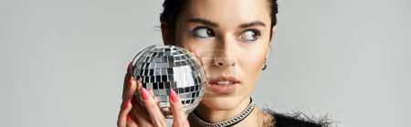 A young woman with short dyed hair poses in stylish attire, holding a disco ball in front of her face.