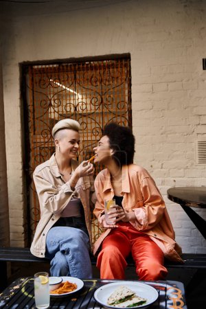 Téléchargez les photos : Un couple diversifié de lesbiennes dégustant un repas ensemble sur un banc. - en image libre de droit