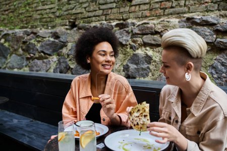 Téléchargez les photos : Deux femmes belles et diversifiées s'assoient à une table, mangeant joyeusement une pizza dans un café confortable. - en image libre de droit
