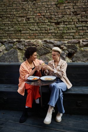Two women, a diverse lesbian couple, enjoy a meal together on a bench in a cafe.