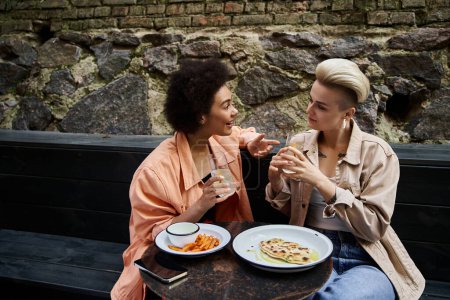 Ein vielseitiges Paar schöner Lesben sitzt an einem Tisch mit Tellern mit leckerem Essen in einem Café.