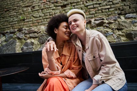 Two diverse, beautiful women sit closely on a bench during their cafe date.