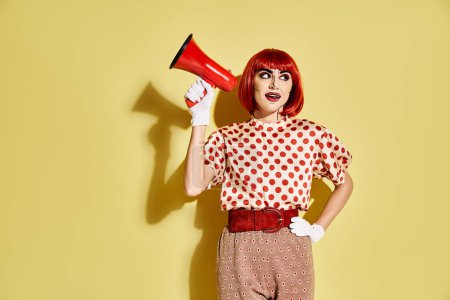 A pretty redhead woman with creative pop art makeup holds a red and white megaphone against a yellow background.