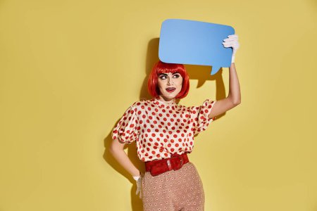 A creative redhead woman with pop art makeup holds a blue speech bubble on a yellow background.