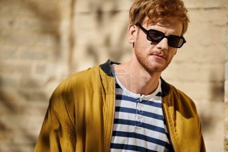 Stylish young man with red hair striding through city streets in a yellow jacket and sunglasses.