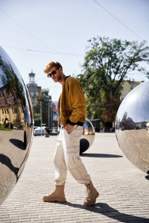 A young red-haired man in debonair attire strolling past two shiny spheres.