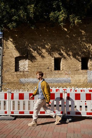 A young red-haired man in debonair attire walking down a city street next to a fence.