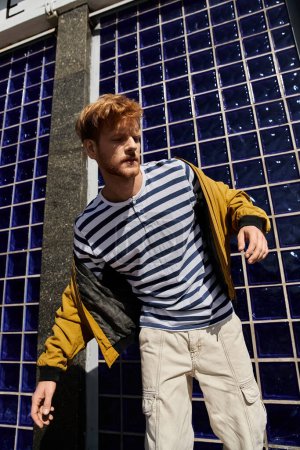 A young red-haired man in debonair attire stands confidently in front of a blue tiled wall.