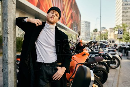 A red-haired man in debonair attire leans against a pole next to a motorcycle.