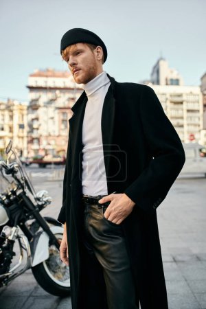 A young red-haired man in debonair attire standing next to a motorcycle in a city parking lot.
