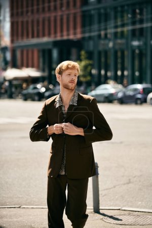 A red-haired, young man stands confidently on a city street in debonair attire.