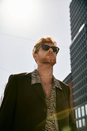 Elegante hombre de pelo rojo con traje y gafas de sol fuera de un edificio imponente.