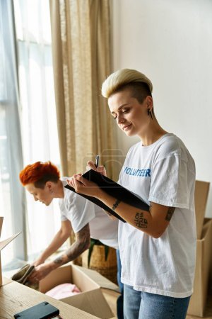A young lesbian couple in matching volunteer t-shirts organizing boxes in a room for charity work.