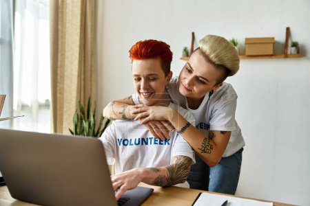 A young lesbian couple in volunteer t-shirts sit together, working on a laptop for charity.