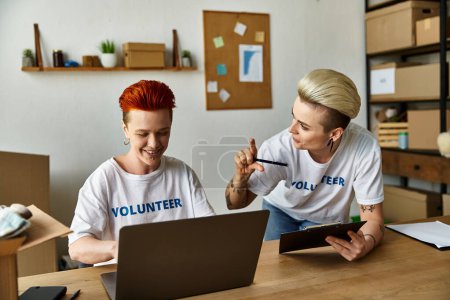 Téléchargez les photos : Deux femmes en t-shirts bénévoles travaillant ensemble sur un ordinateur portable à une table. - en image libre de droit