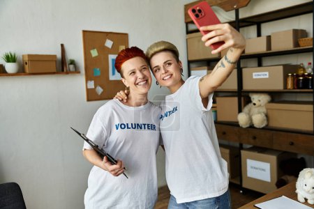 Two women, part of a young lesbian couple, take a selfie in a room as they volunteer together in matching charity t-shirts.
