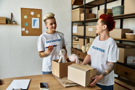 Foto de Joven pareja lesbiana en camisetas voluntarias de pie en la habitación con cajas, encarnando generosidad y compañerismo. - Imagen libre de derechos