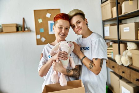 Joven pareja lesbiana en camisetas voluntarias sosteniendo un animal de peluche, difundiendo alegría durante el trabajo de caridad.