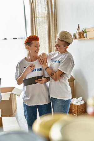 Foto de Una pareja lesbiana con camisetas voluntarias, de pie al lado del otro, participando activamente en el trabajo de caridad juntos. - Imagen libre de derechos
