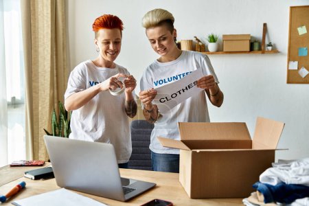 Jeune couple de lesbiennes en t-shirts bénévoles debout devant un ordinateur portable, travaillant ensemble pour une cause de charité.