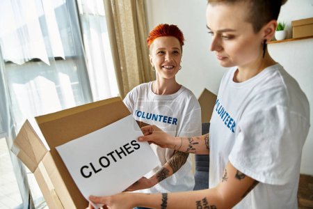 Una joven pareja lesbiana con camisetas voluntarias sosteniendo una caja de ropa donada.