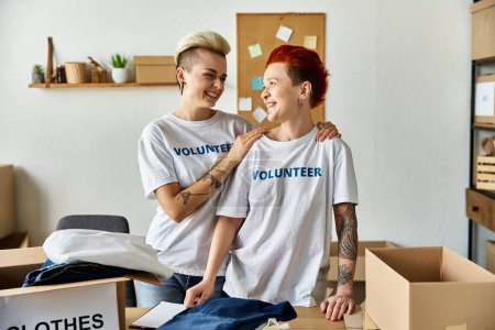 Junges lesbisches Paar in freiwilligen T-Shirts steht vereint in einem Raum, während es Charity-Arbeit verrichtet.