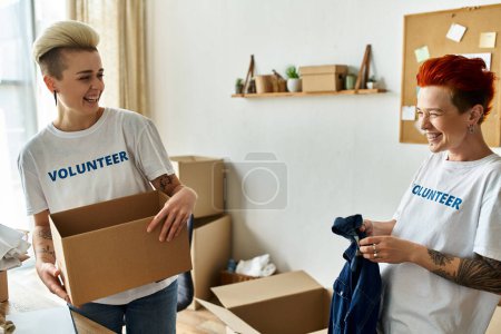 Un par de mujeres con camisetas voluntarias participan en obras de caridad, de pie juntas en una habitación.