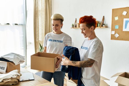 Photo for A young lesbian couple wearing volunteer t-shirts unpack a box of clothes together, engaged in charity work. - Royalty Free Image