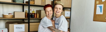 A young lesbian couple wearing volunteer t-shirts stands together in a room, working on a charitable project.