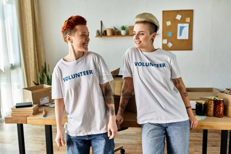 A young lesbian couple in volunteer t-shirts standing together, actively engaging in charity work.