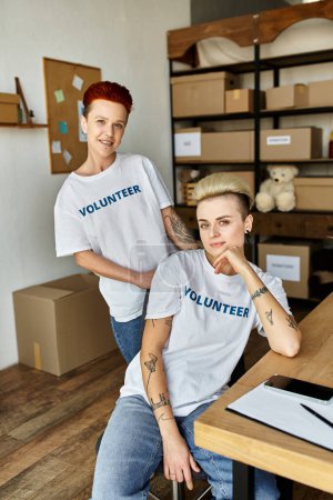 women both wearing volunteer t-shirts, sit at a table engaged in charity work together.