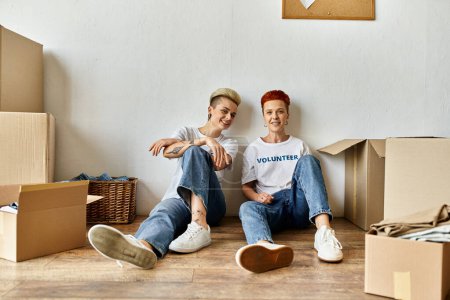 A young lesbian couple in volunteer shirts sit on the floor amid numerous boxes, engaged in charity work together.