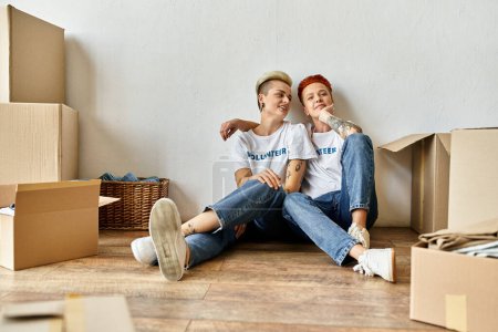 A young lesbian couple in volunteer t-shirts sit together on the floor, engaged in charity work.