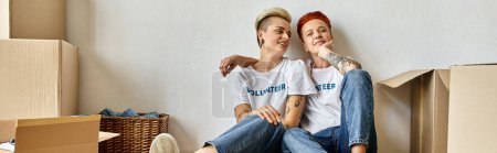 A young lesbian couple in volunteer t-shirts sitting on cardboard boxes, united in charity work.