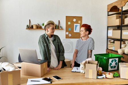A young lesbian couple stands in a room filled with boxes, united in charity work.