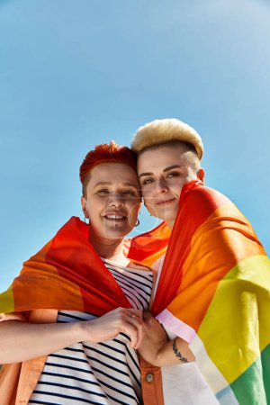 A young lesbian couple hugs tightly, standing proudly with a rainbow flag in an outdoor setting.