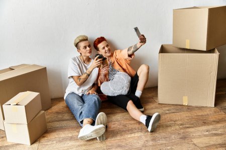 a lesbian couple, are sitting on the floor, snapping a selfie together in a volunteer center surrounded by boxes for charity.