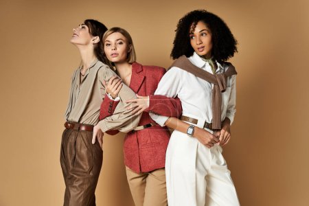 Three beautiful multicultural women in stylish attire pose together on a beige background, showcasing diversity and friendship.