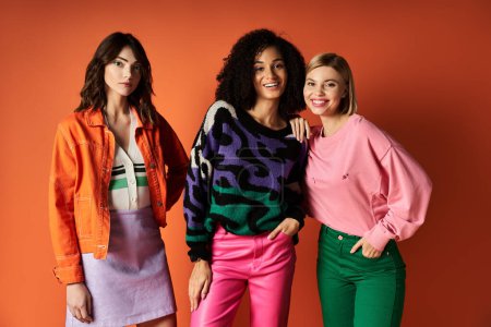 Foto de Tres mujeres jóvenes y hermosas con ropa elegante y vibrante posan juntas sobre un fondo naranja, mostrando diversidad en las culturas. - Imagen libre de derechos