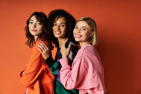 Young, beautiful multicultural women pose in stylish clothes on orange background, showcasing diversity and friendship.