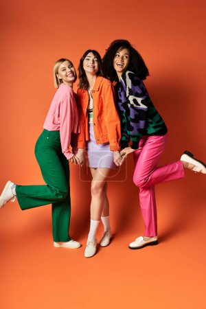 Tres mujeres jóvenes y elegantes posan juntas con un atuendo vibrante sobre un fondo naranja, celebrando la diversidad.